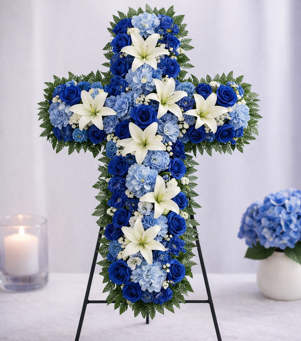 Floral cross arrangement with blue and white flowers on a stand, accompanied by a candle and small vase of flowers.