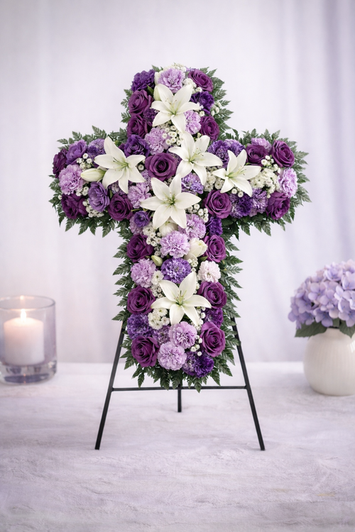 Floral cross arrangement with purple and white flowers on a stand, surrounded by candles and a vase of flowers.